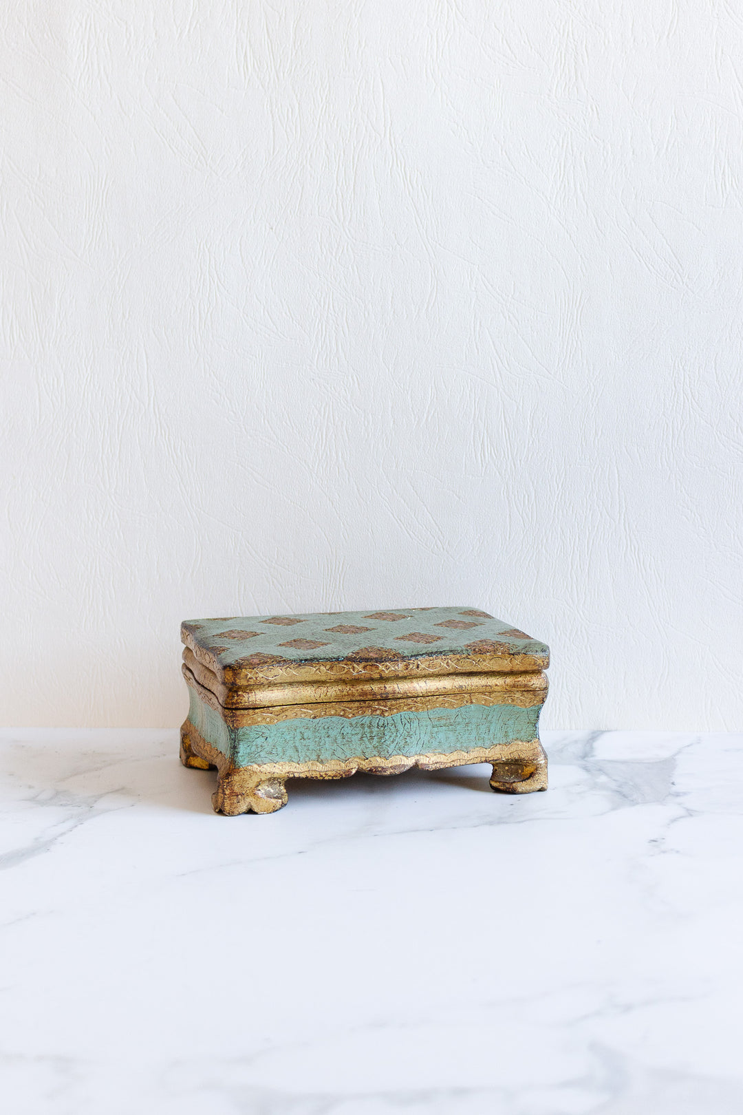 A florentine box with gold and turquoise accents and delicate, vintage design sits on a white marble surface against a textured, light-colored wall.