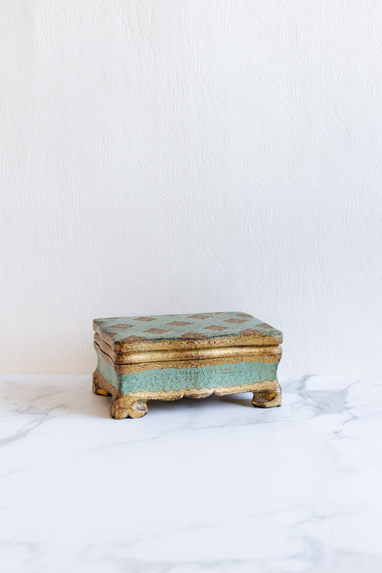 A florentine box with gold and turquoise accents and delicate, vintage design sits on a white marble surface against a textured, light-colored wall.