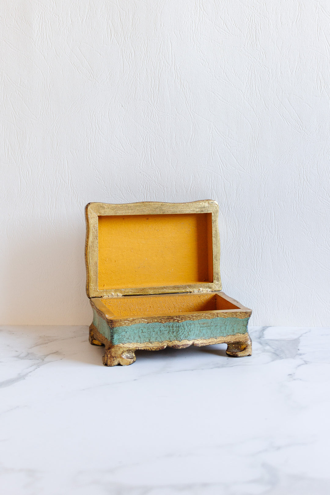 A small florentine box with gold trim and greenish sides rests open on a marble surface. Lined with yellow fabric and featuring decorative feet, it adds charm. Please inquire for exact dimensions if needed.