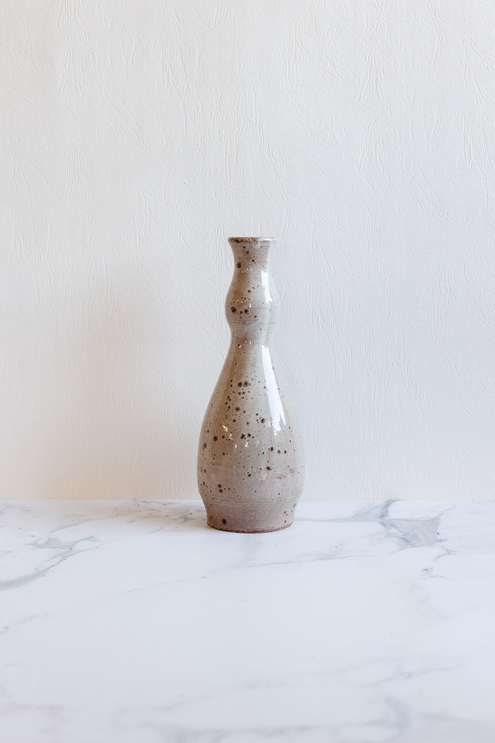 The speckled vase, light gray with elegant dimensions and a narrow neck, sits on a white marble surface against a textured white background.