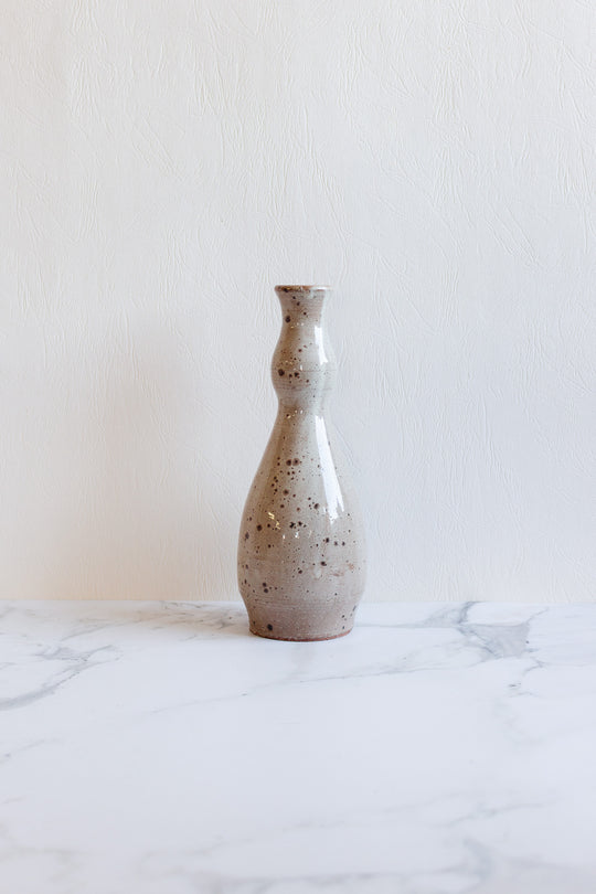 The speckled vase, light gray with elegant dimensions and a narrow neck, sits on a white marble surface against a textured white background.