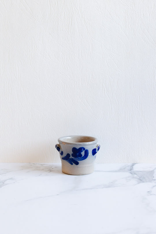 A small, light gray ceramic salt cellar with blue floral designs and compact dimensions sits on a white marble surface against a textured white wall.