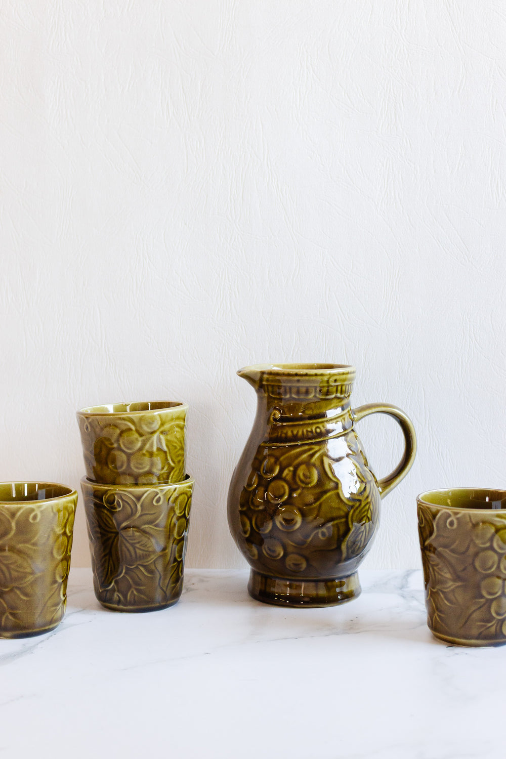 A drinkware set featuring five green ceramic cups and a coordinating patterned pitcher is displayed on a white marble surface with a light textured background.