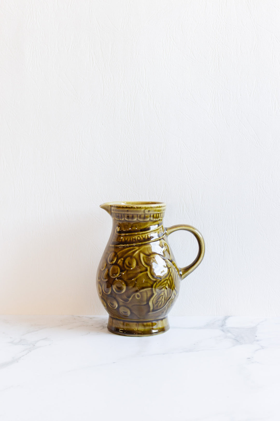 A drinkware set featuring a greenish-brown ceramic pitcher with a floral pattern and elegant contours rests on a white marble surface against a plain background.