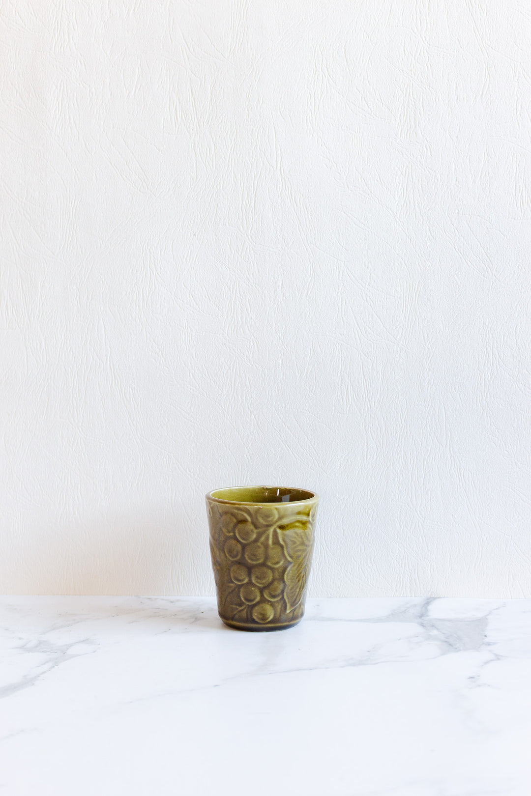 A compact, olive-green ceramic cup from the drinkware set with a textured circular pattern rests on a white marble surface against a white textured wall.