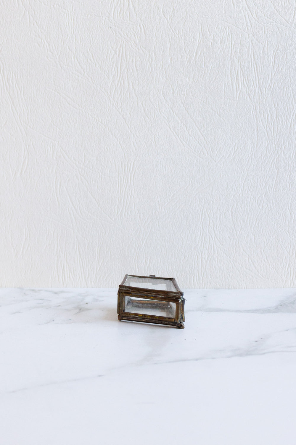 A compact, rectangular pill box with a metal frame and glass sides sits on a white marble surface against a textured white wall.