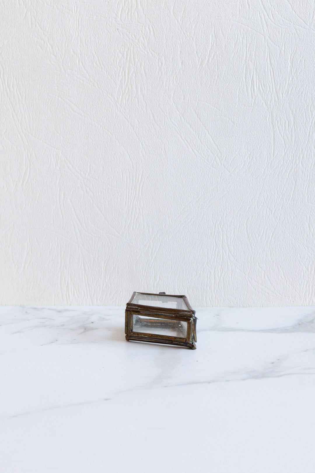 A compact, rectangular pill box with a metal frame and glass sides sits on a white marble surface against a textured white wall.