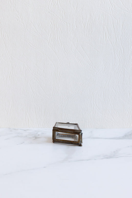 A compact, rectangular pill box with a metal frame and glass sides sits on a white marble surface against a textured white wall.