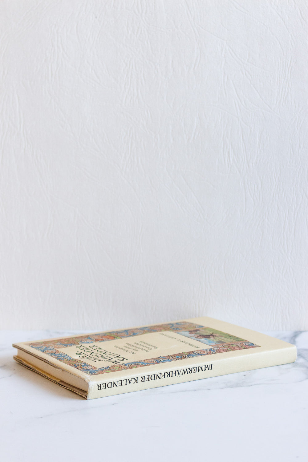 A closed book lies flat on a white marble surface near a textured white wall. Its colorful illustrated cover and distinct dimensions highlight the clean lines, with the partially visible, upside-down title adding visual interest.