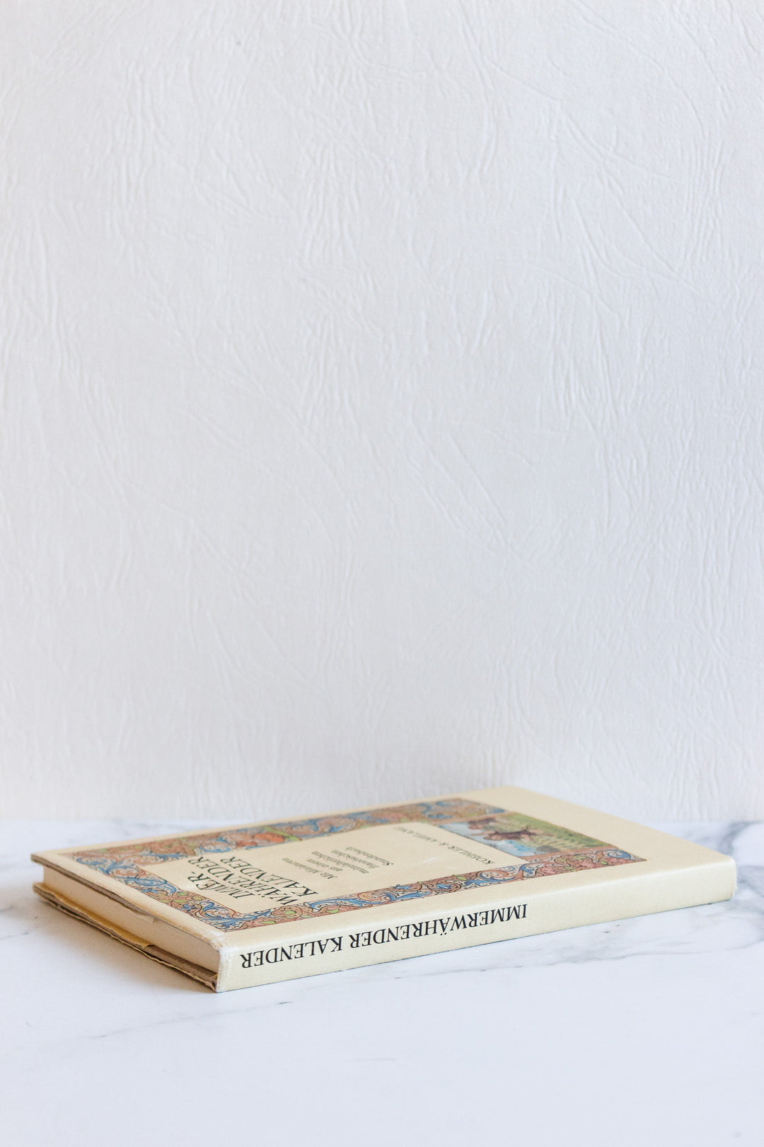 A closed book lies flat on a white marble surface near a textured white wall. Its colorful illustrated cover and distinct dimensions highlight the clean lines, with the partially visible, upside-down title adding visual interest.