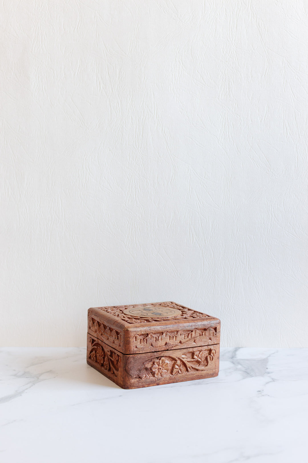 The wood box, featuring intricate carvings and a compact design, is displayed on a white marble surface with a textured white background.