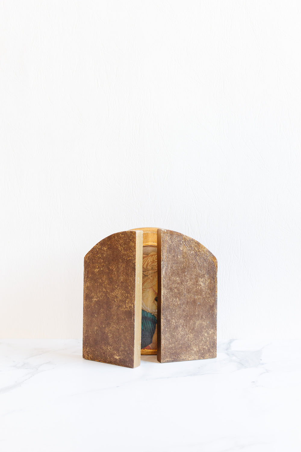 A small, rustic "religious thing" wooden triptych, charmingly sized and partially open to display a painted interior, sits on a white background with a light marble surface.