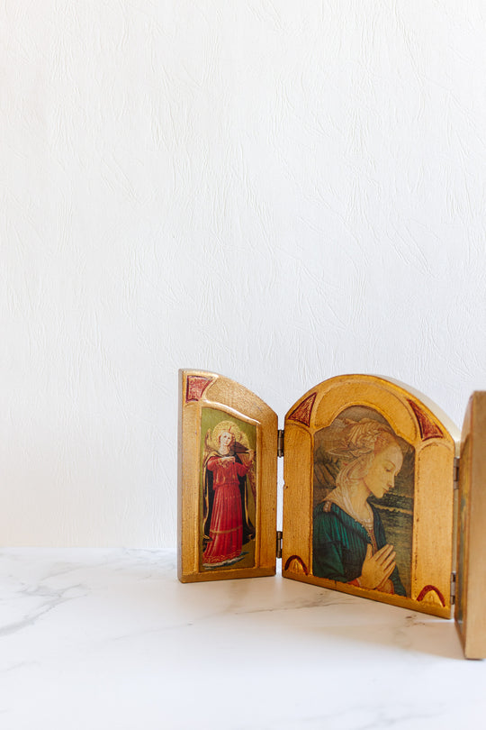 The religious thing is a small, hinged triptych with two painted saintly figures and elegant proportions, displayed on a white marble surface against a light textured wall.