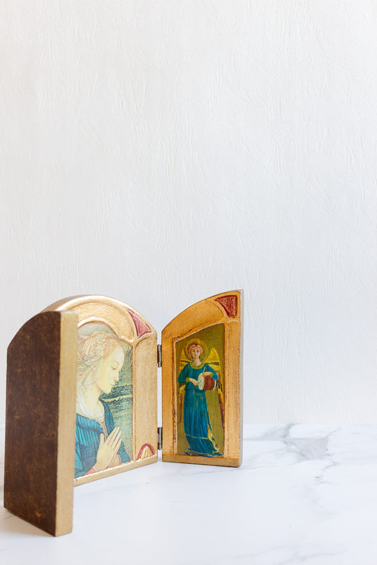 The religious thing, a small wooden diptych with painted icons of a praying woman and a blue-robed figure, is shown open on white marble before a textured white wall, drawing attention to its delicate size.