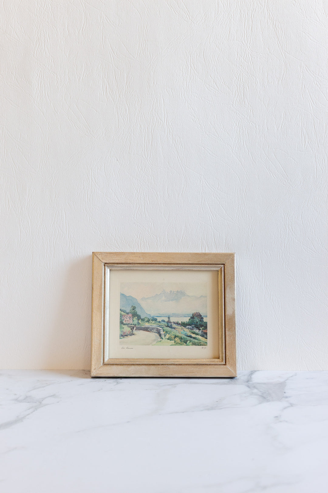 A small art piece with a gold frame, depicting mountains and buildings, sits on a marble surface against a textured white wall—its compact size makes it ideal for any space.