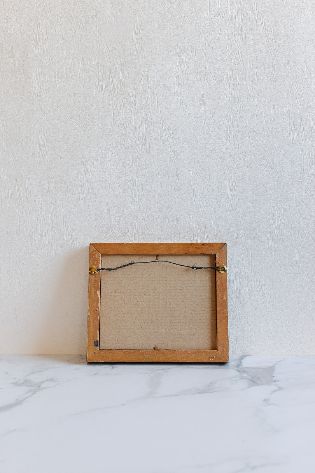 A small empty wooden art frame with visible dimensions is seen from the back, resting on a white marble surface against a textured white wall.