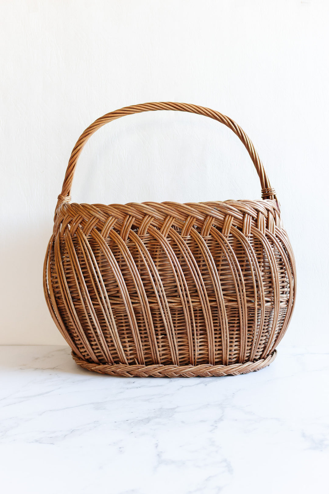 The basket features a round wicker design with a curved handle, displayed on white marble against a plain background, highlighting its simple style and versatile dimensions.