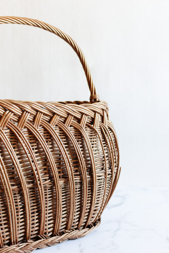 A close-up of the basket with a curved handle on a white marble surface showcases its natural woven texture and highlights its shape against a neutral background.