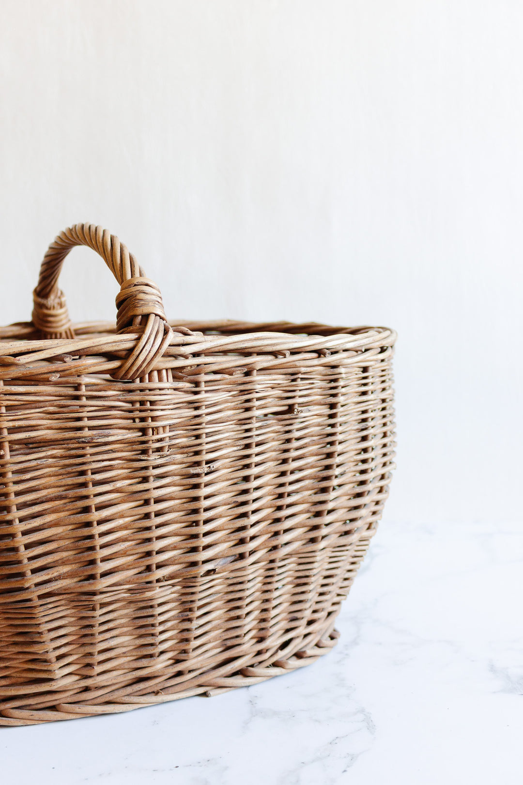 A spacious, empty wicker basket with a curved handle rests on a white marble surface against a light background.
