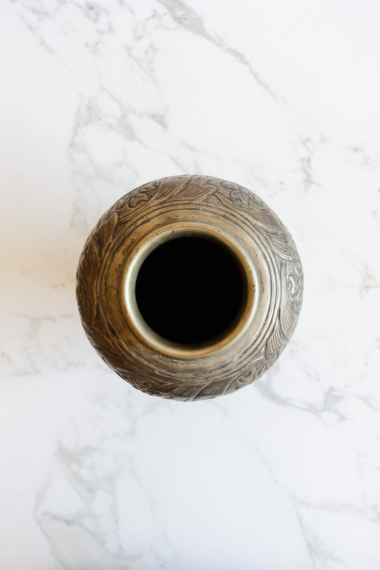 A top-down view of the metal vase shows its intricate engraved patterns and bold dimensions, crafted from brass or bronze and set on a white marble surface. The vase's circular opening appears dark.