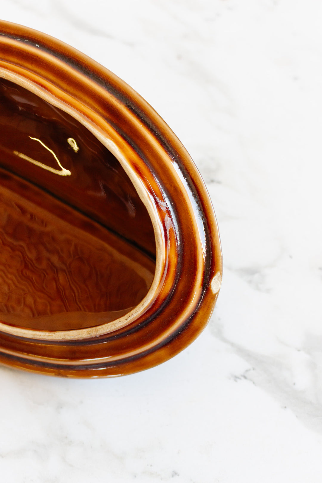Close-up of the fish soupiere, a brown oval ceramic dish with a glossy finish, shown on a white marble surface. The angled top-down view highlights its rim, interior, and dimensions.