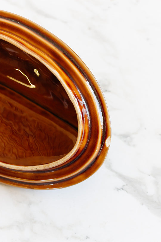 Close-up of the fish soupiere, a brown oval ceramic dish with a glossy finish, shown on a white marble surface. The angled top-down view highlights its rim, interior, and dimensions.