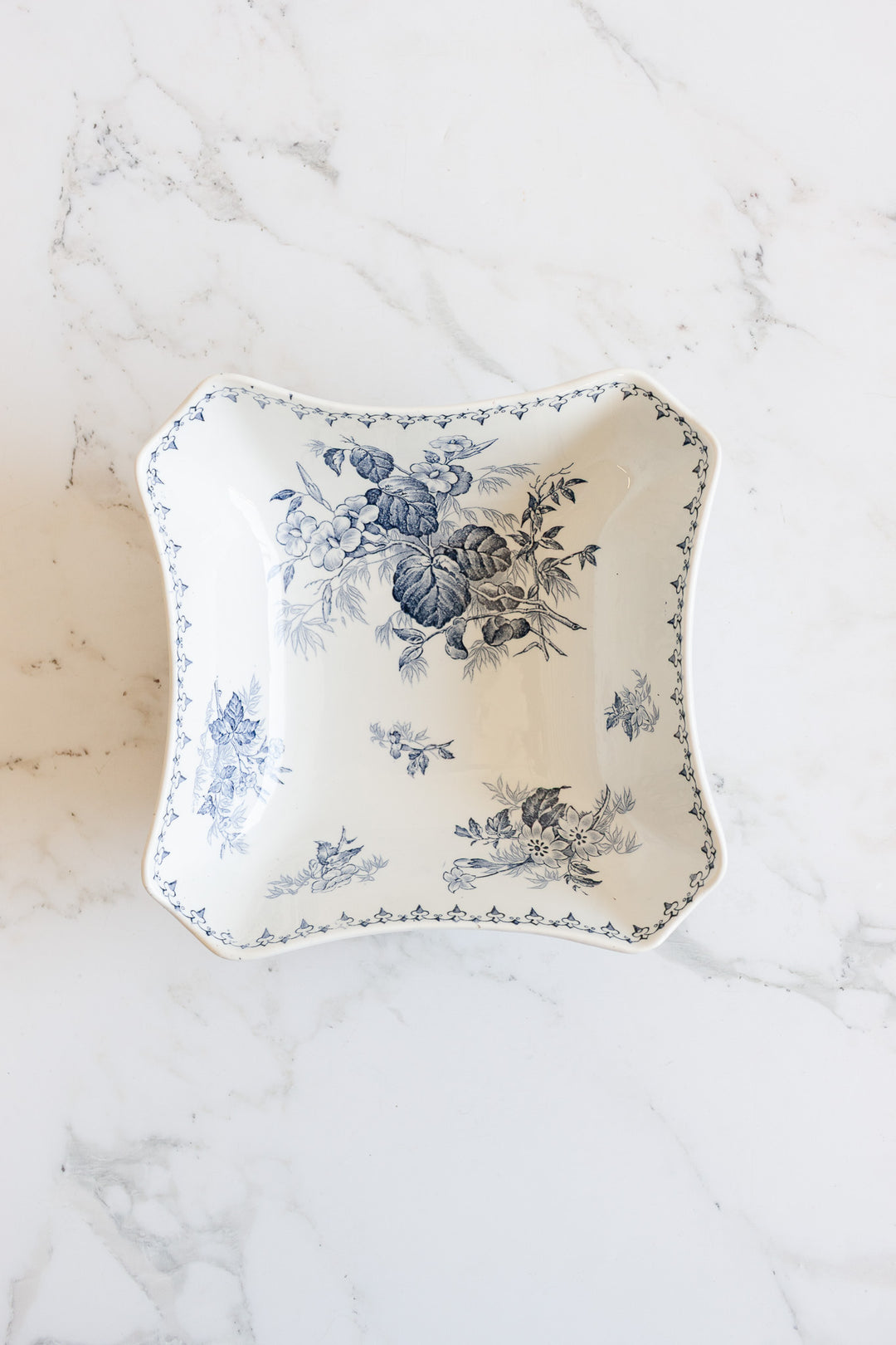 The floral bowl, a square white ceramic piece adorned with blue floral patterns and a decorative border, sits on a white marble surface, showcasing its elegant design.