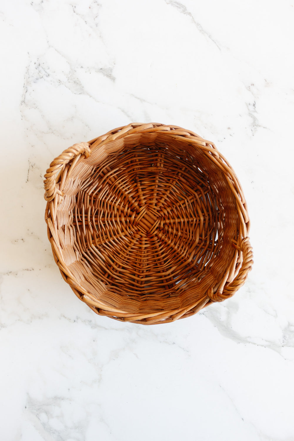 A compact, round woven basket with two handles sits empty on a white marble surface.
