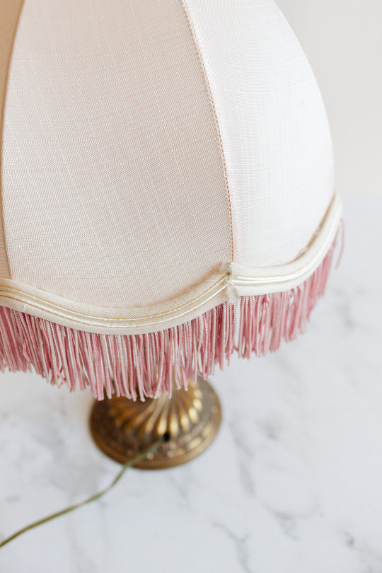 A close-up of the lamp features a cream fabric shade with pink fringe trim, an ornate brass base, and a green power cord on a marble surface, highlighting its vintage style and unique dimensions.