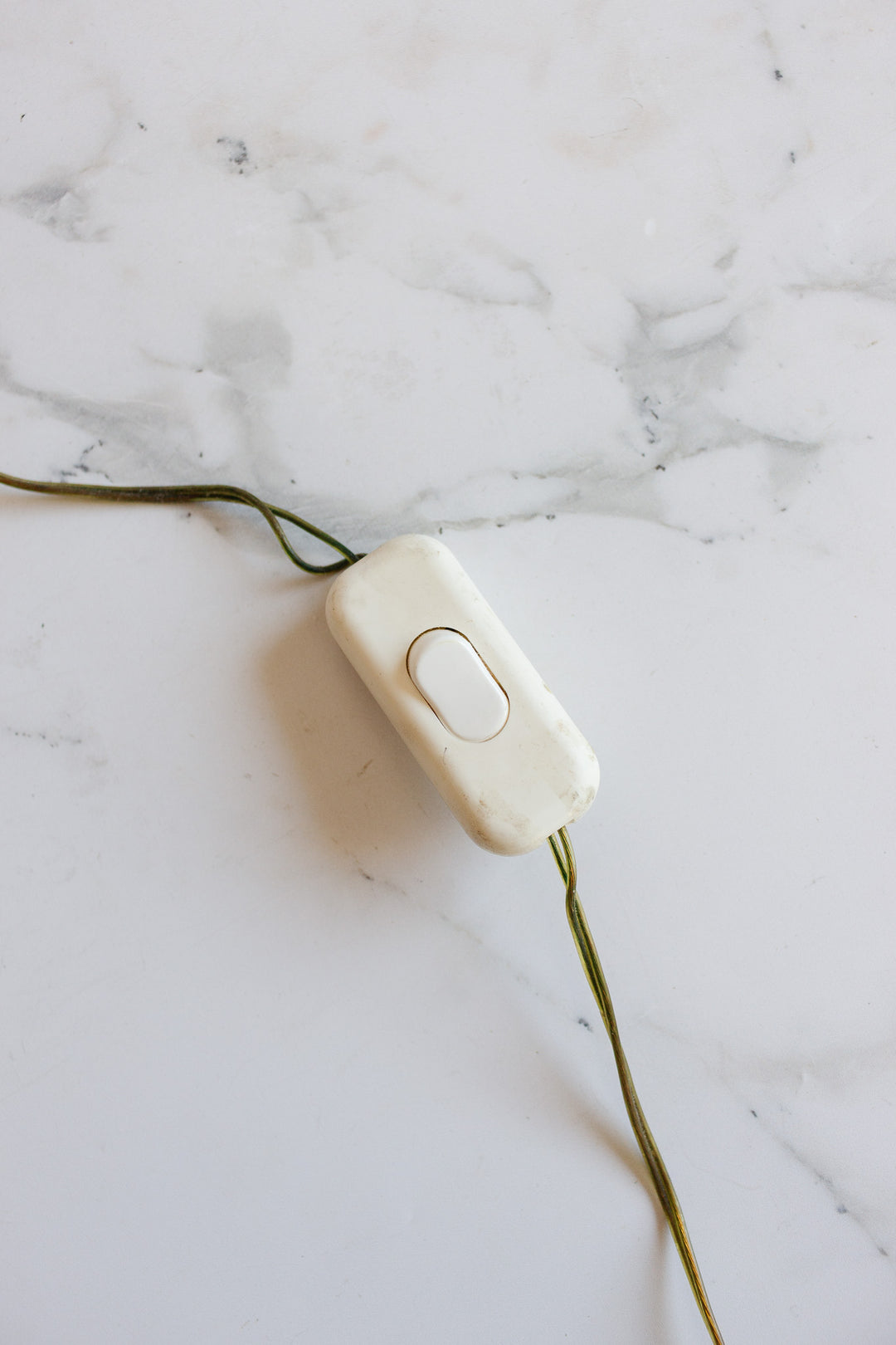 A compact white rectangular inline switch with a central button is connected to the green cord of a lamp and placed on a white marble surface.