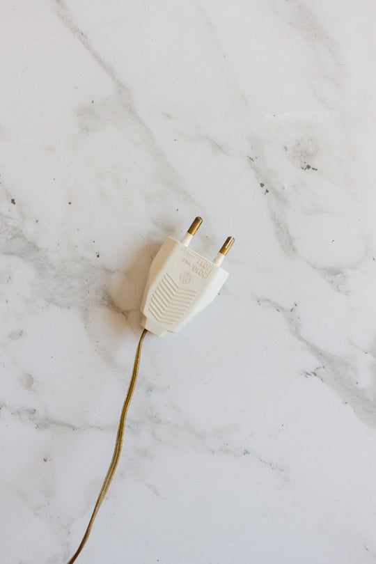 A white two-prong plug with a yellowish-brown cord, part of a compact lamp, rests on a white marble surface with gray veining, making it suitable for various spaces.