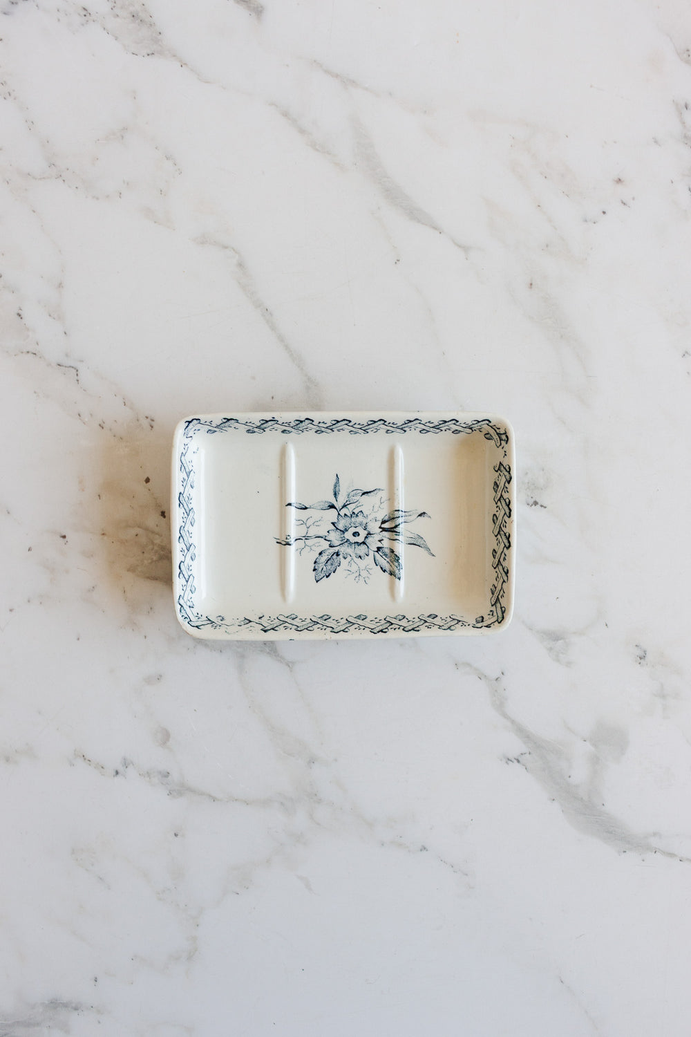 A rectangular ceramic soap holder with a blue floral pattern and decorative border sits on a white marble surface. It features three grooves, a slightly raised edge, and elegant dimensions perfect for serving or display.