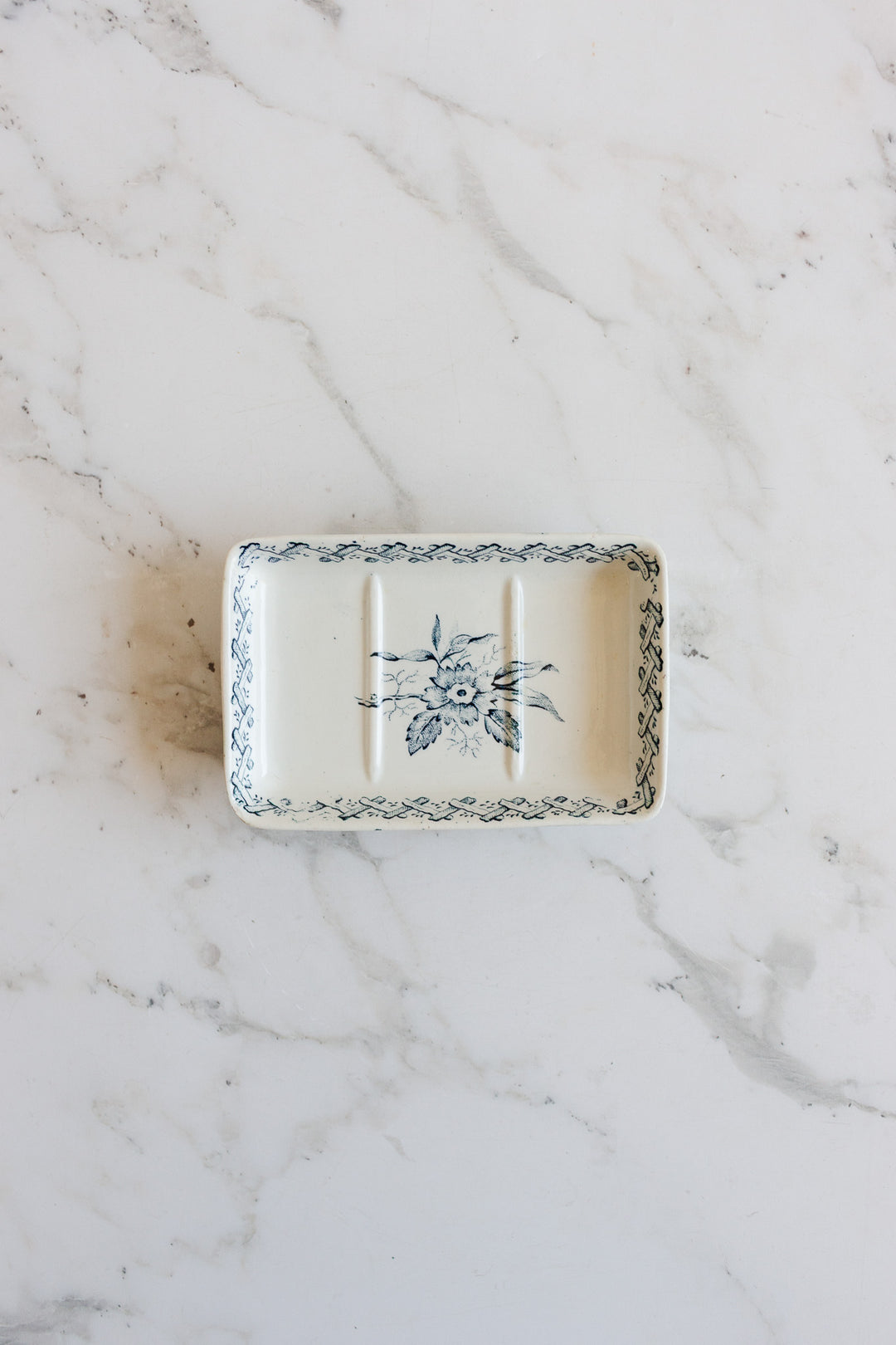A rectangular ceramic soap holder with a blue floral pattern and decorative border sits on a white marble surface. It features three grooves, a slightly raised edge, and elegant dimensions perfect for serving or display.