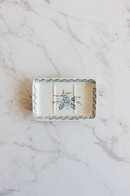 A rectangular ceramic soap holder with a blue floral pattern and decorative border sits on a white marble surface. It features three grooves, a slightly raised edge, and elegant dimensions perfect for serving or display.