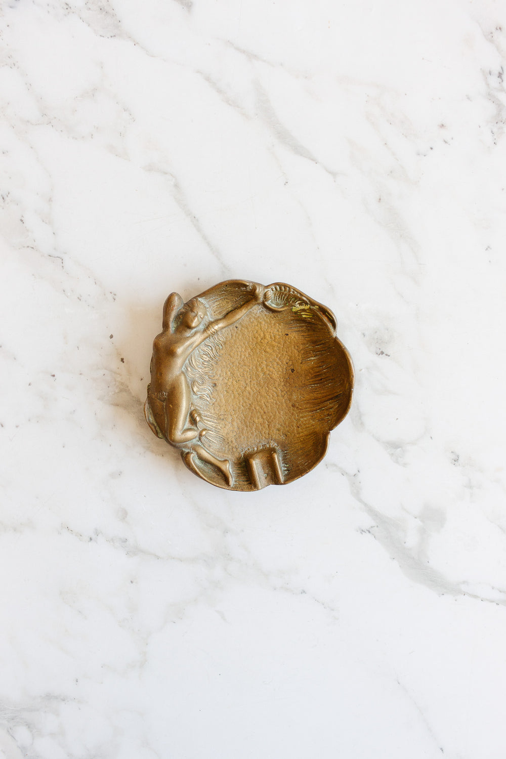 A vintage brass trinket dish shaped like a shell features a raised woman reclining on the left side and is displayed on a white marble surface. Contact us for dimensions.