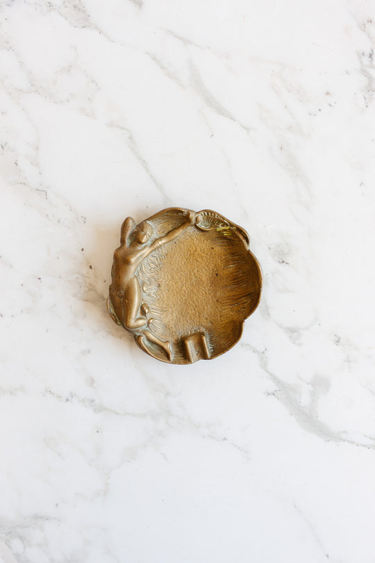 A vintage brass trinket dish shaped like a shell features a raised woman reclining on the left side and is displayed on a white marble surface. Contact us for dimensions.