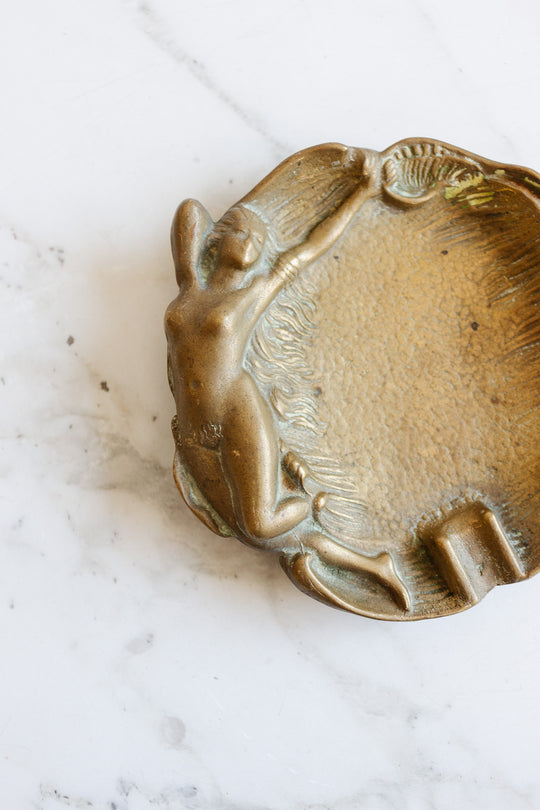 A brass trinket dish featuring an embossed nude woman stretching along the edge, shown on a white marble surface. Contact us for exact dimensions.