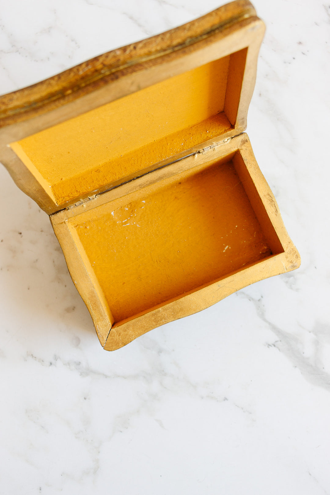 A florentine box with a rectangular shape, rounded edges, and a yellow painted finish inside and out sits open on a white marble surface. The lid is propped up, showcasing its precise craftsmanship.