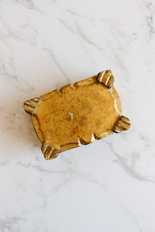 A florentine box with a small, rectangular gold design and four ornate corner feet sits on a white marble surface. Its vintage appeal is highlighted by visible scratches and marks that add to its character.