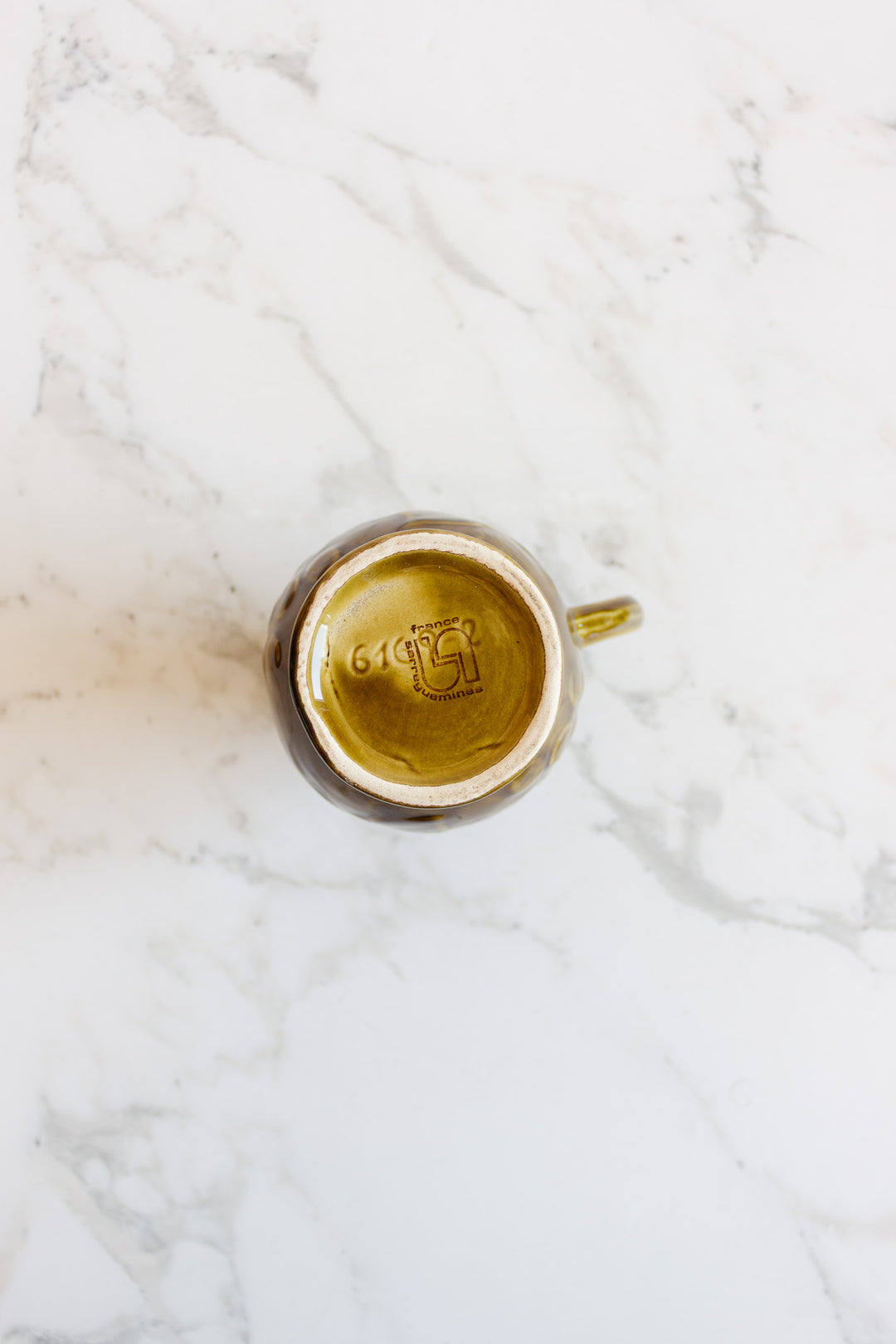 A brown ceramic mug from the drinkware set is upside down on a white marble surface, showing its maker’s mark, dimensions, and the number 614 on the base.