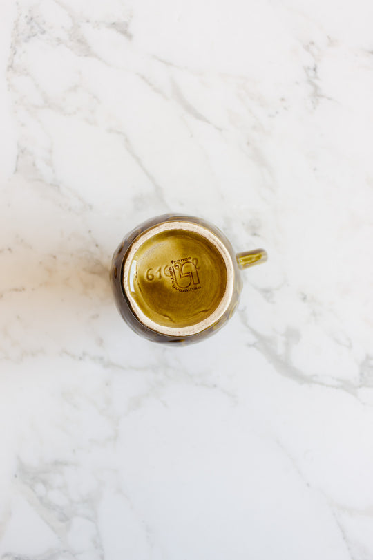 A brown ceramic mug from the drinkware set is upside down on a white marble surface, showing its maker’s mark, dimensions, and the number 614 on the base.