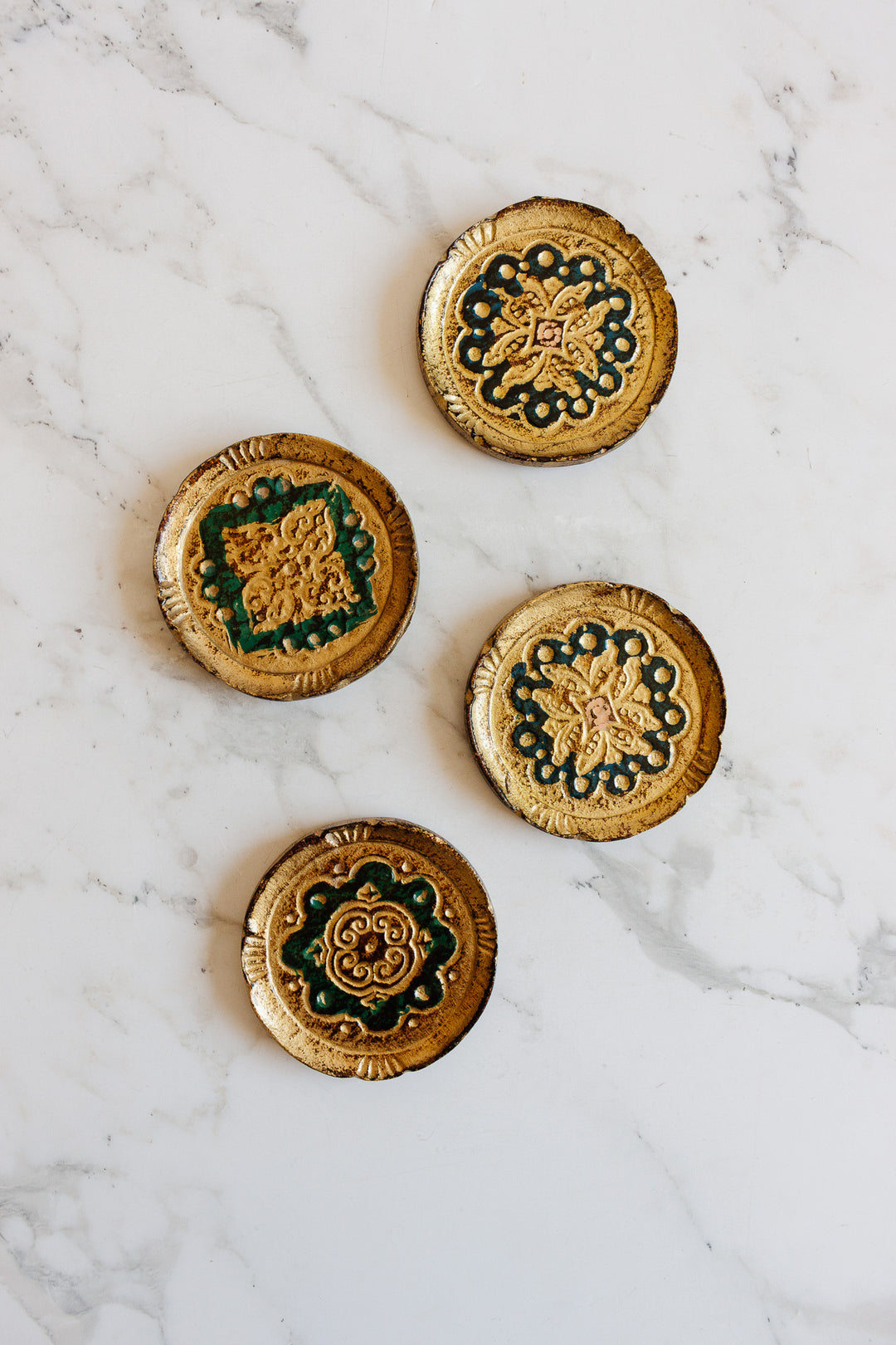 Four ornate florentine coasters, featuring round and square shapes with gold and green embossed designs, are displayed on a white marble surface. Their intricate patterns, textures, and sizes give each one a unique touch of elegance.