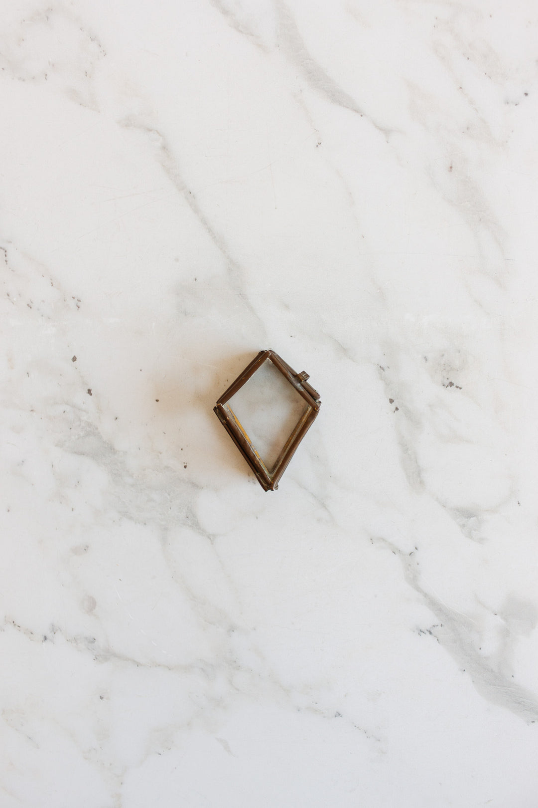 A compact, diamond-shaped metal pill box with sleek dimensions rests on a white marble surface with gray veining.