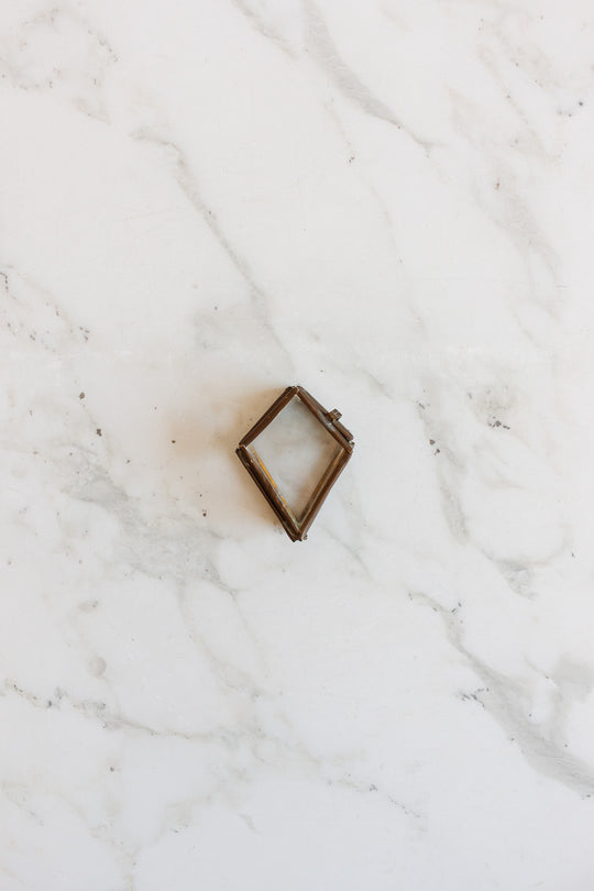 A compact, diamond-shaped metal pill box with sleek dimensions rests on a white marble surface with gray veining.