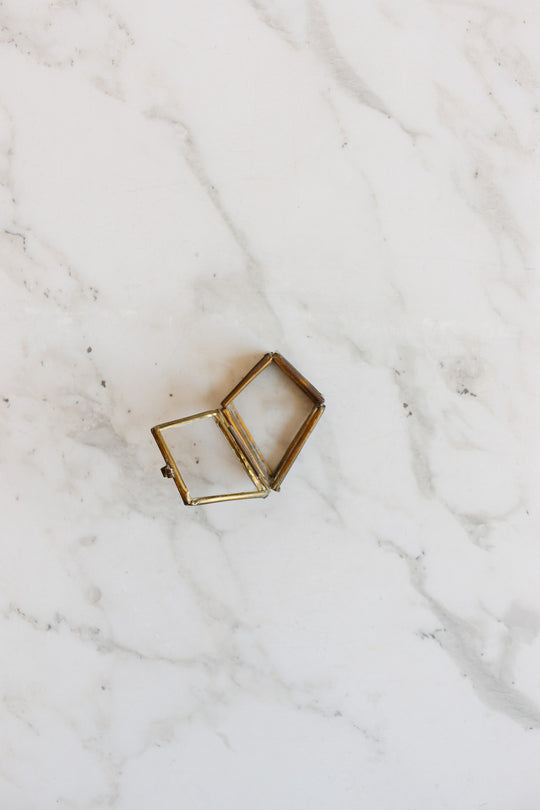 A small, empty pill box crafted from glass and brass, with elegant dimensions, sits open on a white marble surface with gray veining.