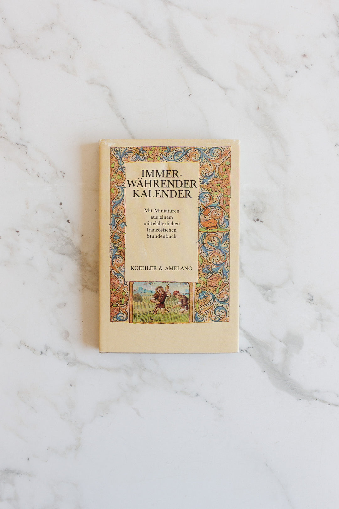 A beige book titled "Immerwährender Kalender" with a colorful ornate border and medieval-style cover illustration rests on a white marble surface, its elegant design highlighted by the harmonious dimensions.