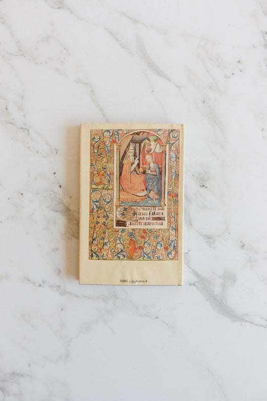 A vintage book with an ornate, colorful medieval illustration on the cover and worn edges sits on white marble. Intricate patterns frame the image, and its unique dimensions highlight the timeless charm of this book.