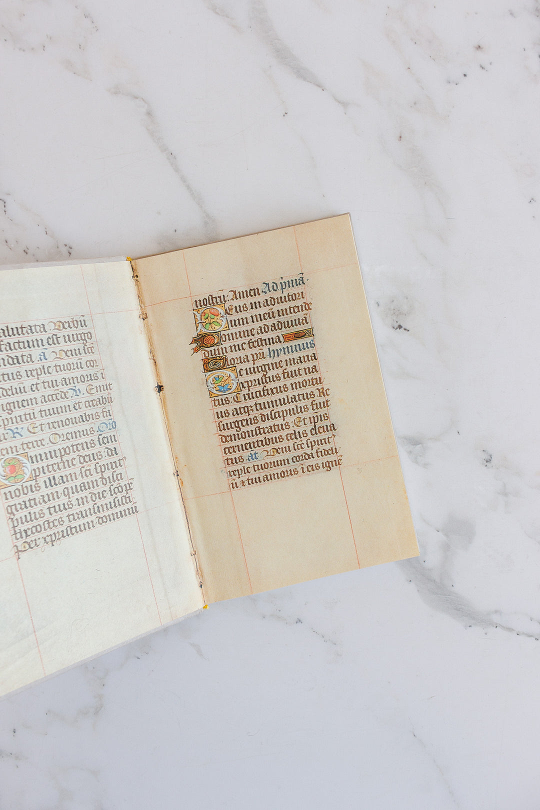 A large open book rests on marble, showcasing decorative gold accents, an ornate initial letter, and neat handwritten Latin text on aged parchment.