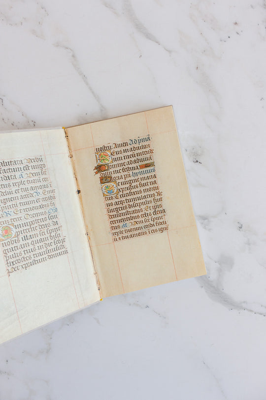 A large open book rests on marble, showcasing decorative gold accents, an ornate initial letter, and neat handwritten Latin text on aged parchment.