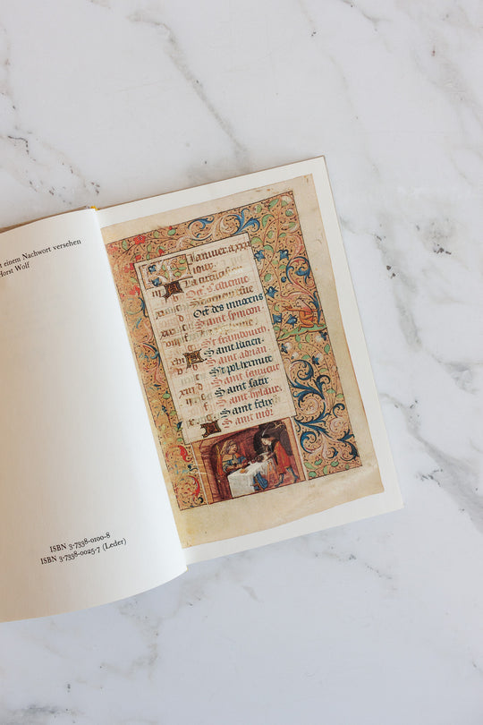 An open book lies on a marble surface, displaying a page with ornate medieval-style calligraphy and colorful floral designs, next to a blank page listing the ISBN numbers and dimensions at the bottom.